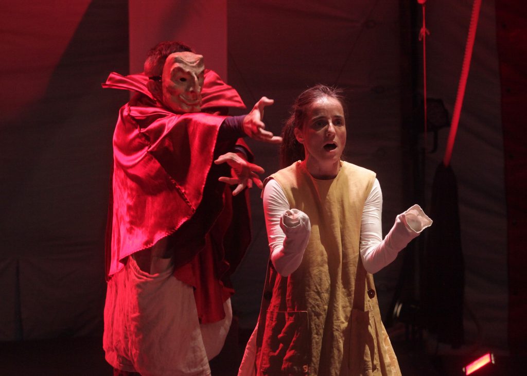 Výjav z divadelnej hry: Osoba v maske a červenom plášti sa naťahuje po žene