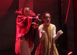 Výjav z divadelnej hry: Osoba v maske a červenom plášti sa naťahuje po žene