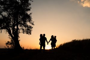 Silueta rodiny pri západe Slnka. Mama a otec sa držia za ruky. Na rukách majú deti.