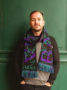 Muž v strednom veku so šálom okolo krku pózuje pri zelenej stene