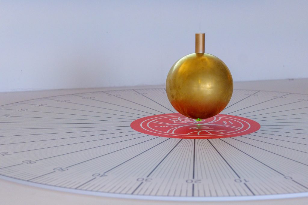 Detail zlatej gule Foucaultovho kyvadla nad stupnicou