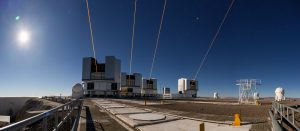 Budova astronomického observatória počas jasného dňa a štyri laserové lúče smerujúce k oblohe