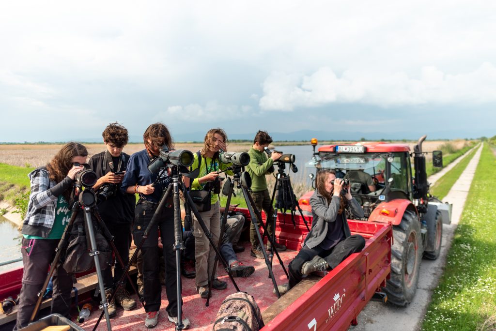 Skupina ľudí stojí na vlečke traktora a pozoruje vtáky pomocou ďalekohľadov