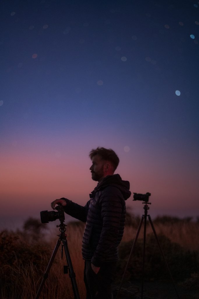 Fotograf s fotoaparátmi sa pozerá do diaľky počas súmraku
